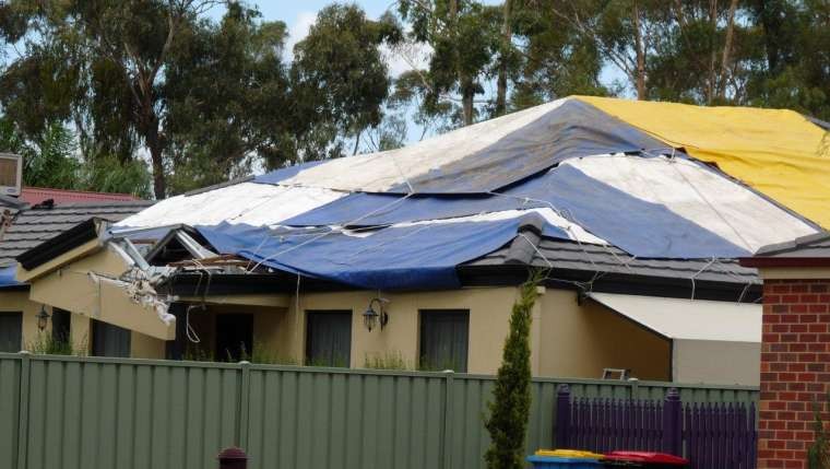 A house's roof showing significant damage caused by storms, covered with multiple tarps.